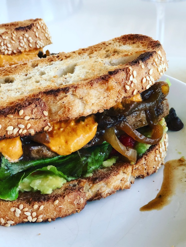 Smokey Tofu Sandwich, vegan plantbased with caramelized onions, oven-roasted veggies,toasted bread, lettuce on white plate with white background